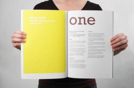 a person in a black shirt holding up a book. On the left-hand side of the book is a yellow page that read "step one" in white font. On the right-hand site is a white page with the word "one" written in large brown font.